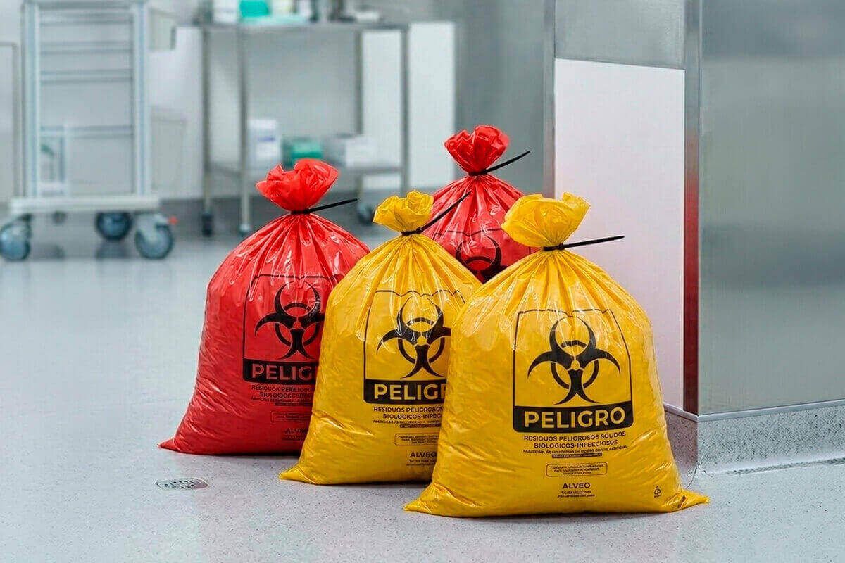 Manos con guantes acomodando bolsas RPBI amarillas y rojas dentro de contenedores etiquetados en un cuarto de almacenamiento de insumos médicos con luz natural